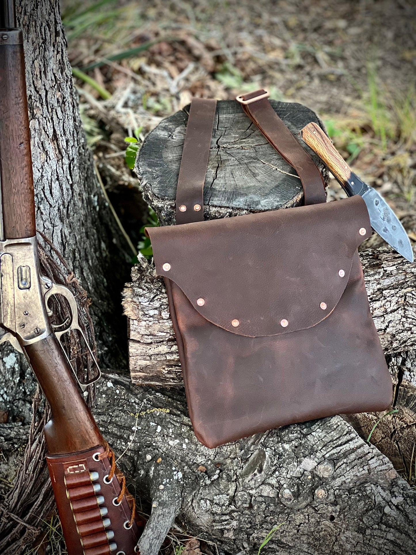 The Culper Haversack made from Kodiak leather, featuring copper hardware and decorative rivets, placed beside a rifle and knife.