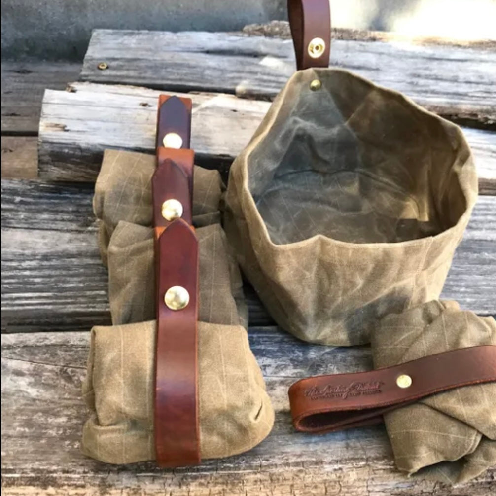 Waxed canvas dog bowl with leather straps, folded and unfolded, on a wooden surface.