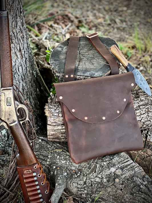 The Culper Haversack made from Kodiak leather, featuring copper hardware and decorative rivets, placed beside a rifle and knife.